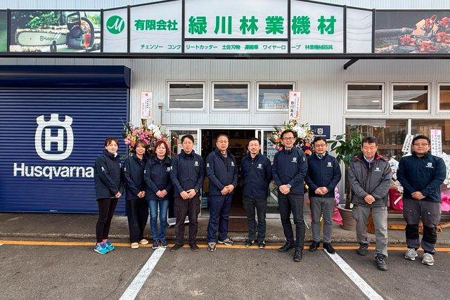 ハスクバーナのブランドと様々な林業・建設機材の看板が掲げられた店の前に立つ人々のグループ。