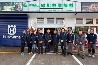 ハスクバーナのブランドと様々な林業・建設機材の看板が掲げられた店の前に立つ人々のグループ。
