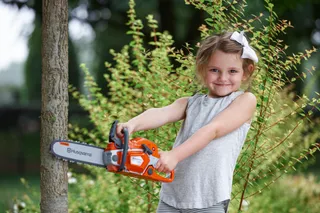 A child holding a Husqvarna toy chainsaw in a garden setting.