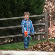 A child walking in a garden with a toy leaf blower near a pile of leaves.