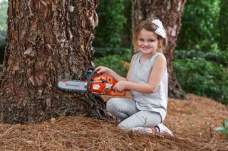 Dziecko klęczące na ziemi, trzymające piłę łańcuchową opartą o pień drzewa w leśnej scenerii.