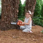 A child kneeling on the ground, holding a chainsaw against a tree trunk in a forest setting.