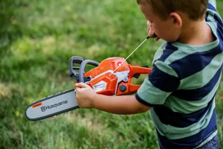 Child holding a Husqvarna toy chainsaw in a grassy area.