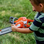 Child holding a Husqvarna toy chainsaw in a grassy area.