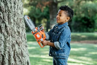 A child holding a toy chainsaw against a tree trunk in a garden setting.