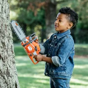 A child holding a toy chainsaw against a tree trunk in a garden setting.