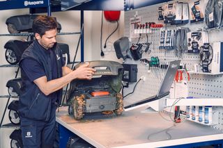 Eine Person, die in einer Werkstattumgebung an einem Mähroboter arbeitet, umgeben von verschiedenen Werkzeugen und Geräten.