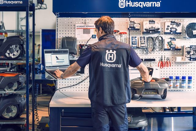 A person in a workshop setting working on a robotic lawn mower with a laptop displaying related information.