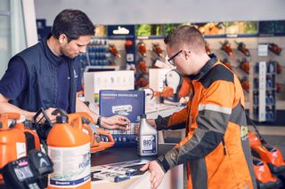 Divi cilvēki veikalā apspriež produktu, ko ieskauj dažādi dārza darbarīki un aprīkojums.