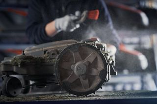 Close-up of a person cleaning a lawn mower blade with a brush in a workshop setting.