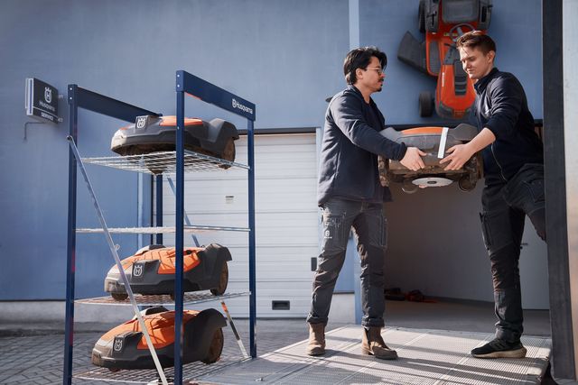 Two people handling a robotic lawn mower near a storage rack with additional mowers.
