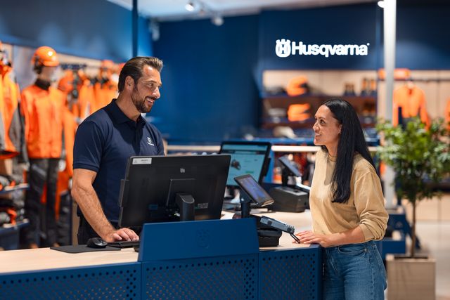 Eine Person, die mit einem Filialmitarbeiter an einer Husqvarna-Verkaufstheke interagiert, während im Hintergrund Produkte ausgestellt werden.