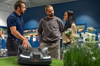 Tre personer diskuterar en robotgräsklippare utställd på konstgräs i ett showroom.