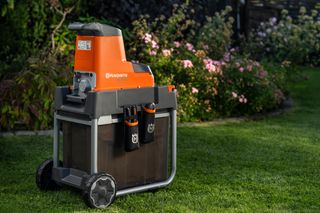 En kompostkvarn från Husqvarna placerad på en gräsmatta med blommande blommor i bakgrunden.