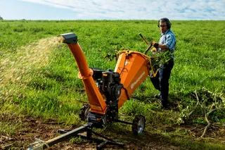 Triturador de Galhos Husqvarna GC 5100PE. Uma pessoa usando um picador de madeira em um campo gramado, com aparas de madeira sendo expelidas da máquina.