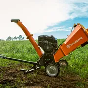 Triturador de Galhos Husqvarna GC 5100PE. Um picador de madeira em um campo, parcialmente visível com rodas e calha.