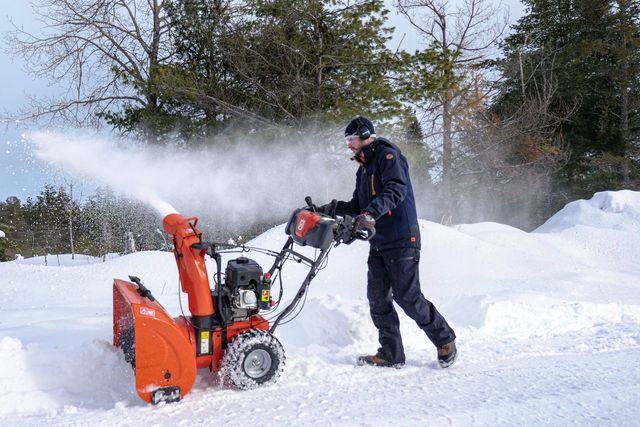 A person operating a snow blower on a snowy path surrounded by trees.