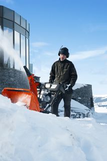 Une personne qui utilise une souffleuse à neige près d’un bâtiment moderne dans un paysage enneigé.