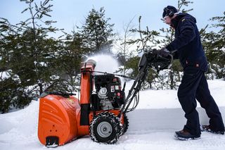 A person operating a snow blower on a snowy path with trees in the background.