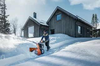 Une personne utilise une souffleuse à neige pour déneiger un chemin devant une maison en bois dans un paysage enneigé.