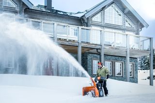 En person som bruker en snøfreser til å rydde snø foran et stort hus.