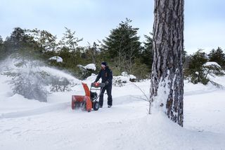 A person using a snow blower to clear snow in a snowy forest setting.