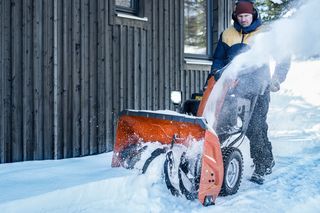 En person som betjener en snøfreser for å rydde snø foran en trebygning.