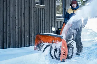 Une personne qui utilise une souffleuse à neige pour déneiger devant un bâtiment en bois.