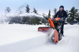 A person operating a snow blower on a snowy path, clearing snow with trees in the background.