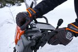 A person wearing Husqvarna gloves, operating a snow blower in a snowy environment.