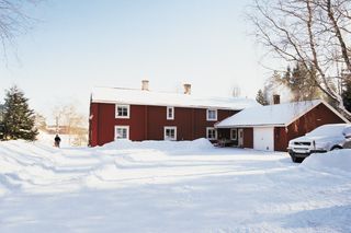Et snødekt landskap med et rødt trehus og et parkert kjøretøy, omgitt av trær.