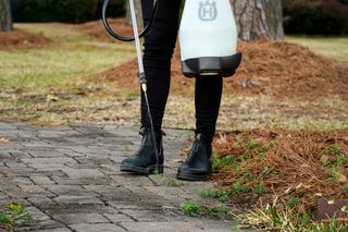 A person using a Husqvarna sprayer on a paved walkway in a garden setting.