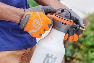 A person holding a Husqvarna spray bottle, wearing gloves in a garden setting.