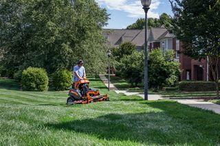 A person mowing a lawn with a ride-on mower in a residential neighborhood.