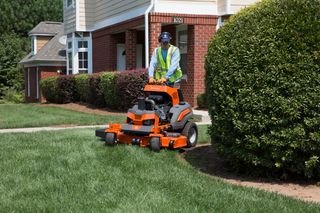 A person mowing lawn with a ride-on mower near a brick house.