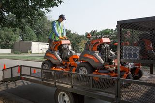 A worker in work gear loading two ride-on mowers onto a trailer.