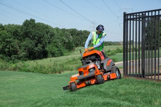 A person operating a ride-on mower on a grassy slope near a metal fence.