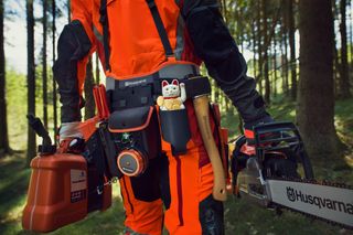 A person in work gear holding a Husqvarna chainsaw and carrying various tools in a forest.