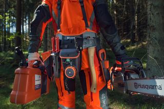 Людина в робочому спорядженні тримає в руках ланцюгову пилку Husqvarna й каністру з пальним, що стоїть у лісі.