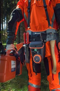 Людина в робочому спорядженні тримає в руках ланцюгову пилку Husqvarna й каністру з пальним, що стоїть у лісі.