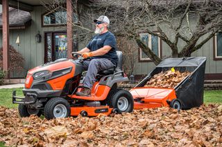 42" Lawn Sweeper. A person using a ride-on mower with a leaf collector attachment to clear fallen leaves in a garden.