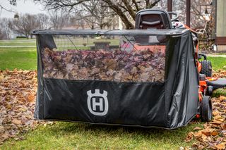 A ride-on mower collecting fallen leaves in a garden, with a visible Husqvarna logo on the leaf bag.