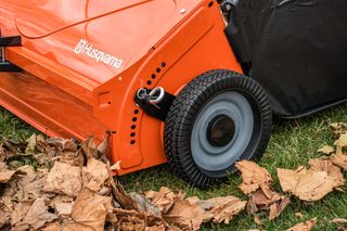 42" Lawn Sweeper. Close-up of a Husqvarna lawn mower wheel on grass with fallen leaves.