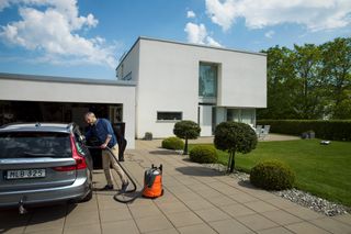 En person som använder en dammsugare för att rengöra en bil som står parkerad framför ett modernt hus.