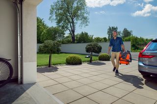 A person carrying a leaf blower in a garden setting, with a parked car nearby.