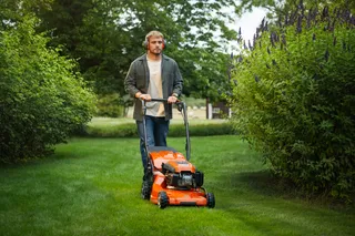 HUSQVARNA LC 347VE. Oseba, ki kosi vrtno trato s kosilnico, ki je hodna za seboj, med grmovjem in drevesi.
