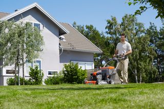A person mowing a lawn with a walk-behind mower in a residential garden.
