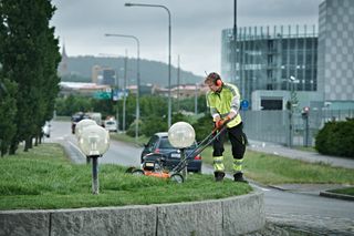 A person using a lawn mower to trim grass on a roadside curb.