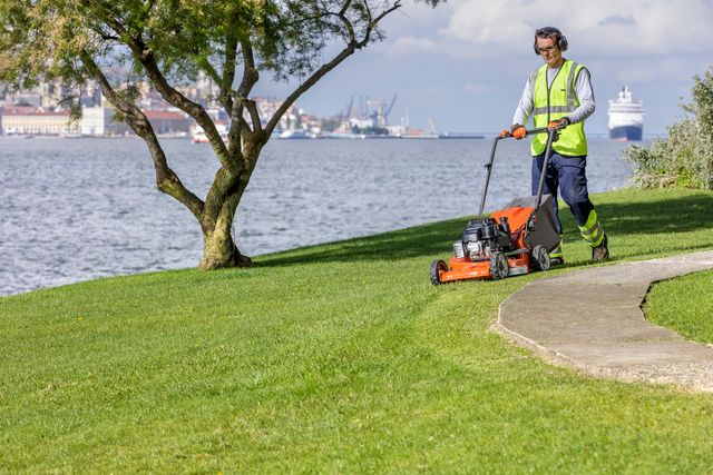 A person mowing a lawn near a waterfront with a push lawn mower.