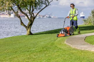 A person mowing a lawn near a waterfront with a push lawn mower.